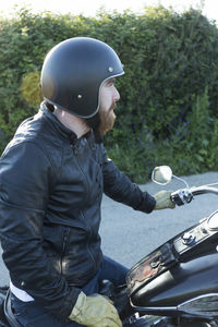 Side view of biker looking away while sitting on motorcycle against plants during sunset