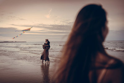 Rear view of woman on beach against sky during sunset with girls playing kite at the background 
