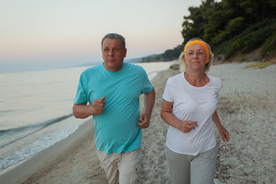 Mature couple jogging at beach against sky during sunset