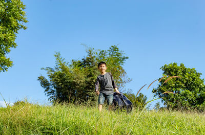 Full length portrait of boy holding backpack while walking on grass against clear sky