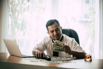 Smiling mid adult businessman holding money and gun while sitting in office