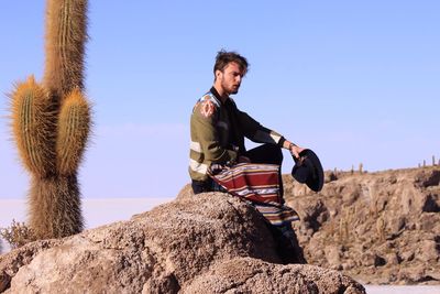 Young man sitting on rock against clear sky