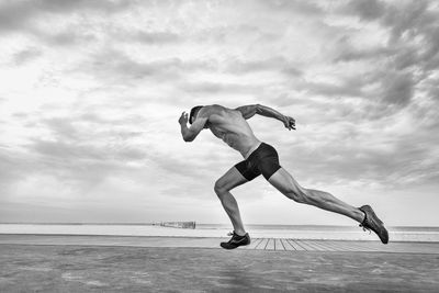 Side view of shirtless man taking running stance against sky