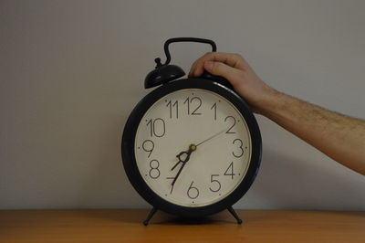 Cropped hand over alarm clock on table against wall