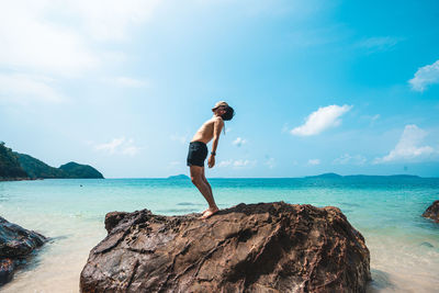 Full length of shirtless man on rocks at sea shore against sky