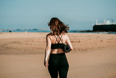 Back view of slim unrecognizable female in sportswear and with yoga mat walking along beach near river on sunny day