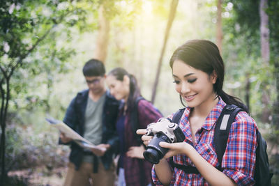 Smiling woman holding camera while friends using map in forest