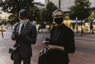 Male and female colleagues standing with smart phone on footpath during pandemic