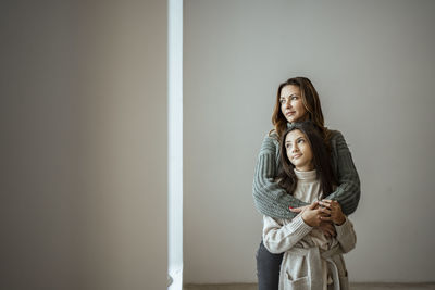 Mother and daughter looking away while embracing each other against wall at home