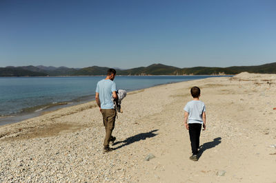 Man and preschool boy in blue identical t-shirts walk side by side on huge deserted beach and sea