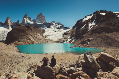 Rear view of man and woman sitting on rock near lake and mountains