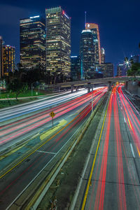 Light trails on city street by buildings against sky at night