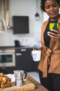 Confident female architect using phone while standing with cup at home