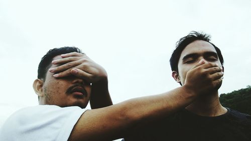 Low angle view of friends covering eyes and mouth against clear sky