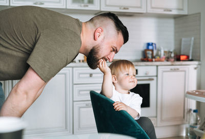 Happy father young man feeding and having fun with baby girl little daughter in kitchen at home