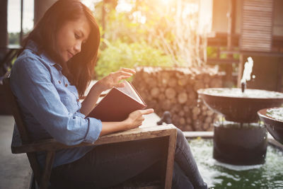 Side view of woman reading book while sitting at yard