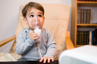 Cute boy inhaling through respiratory mask while sitting on chair at home