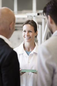 Smiling engineer showing machine part to businessmen in manufacturing plant