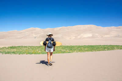 Full length rear view of woman walking with sandboard in desert