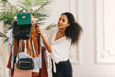 Young woman looking at clothes and purses on rack while standing at home