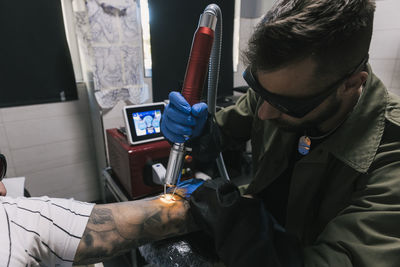 Side view of focused male making laser tattoo on arm of tired shaved cropped unrecognizable client in tattoo salon