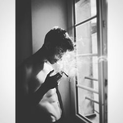 Shirtless man smoking cigarette while standing by window at home