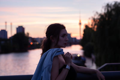 Young woman looking away while standing by railing during sunset