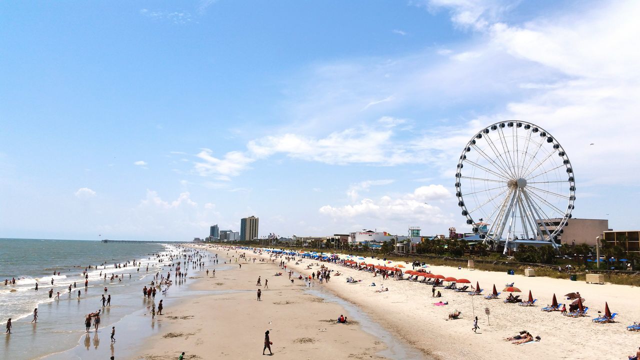 beach, sea, sand, large group of people, sky, outdoors, ferris wheel, leisure activity, vacations, travel destinations, nature, horizon over water, day, amusement park, water, people