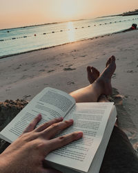 Low section of woman reading book at beach against sky