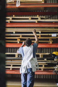 Rear view of male carpenter searching for wooden plank on pallet stack in warehouse