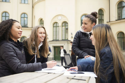Multi-ethnic female friends smiling while studying at table in schoolyard