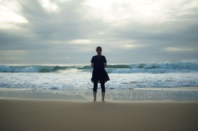 Rear view of man standing at seashore against cloudy sky