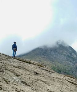 Rear view of hiker walking on rock by mountain against cloudy sky