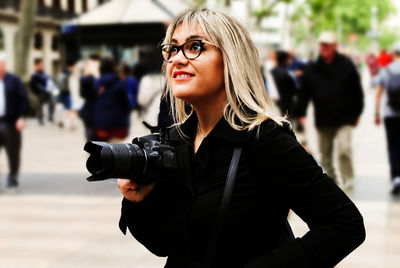 Portrait of young blonde woman, wearing glasses and holding camera in the hand, while looking away.