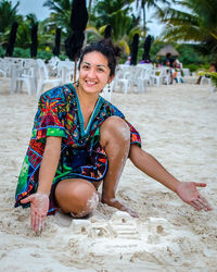Portrait of smiling young woman sitting by sandcastle on beach