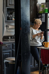 Smiling mid adult woman text messaging on smart phone while standing in kitchen at home