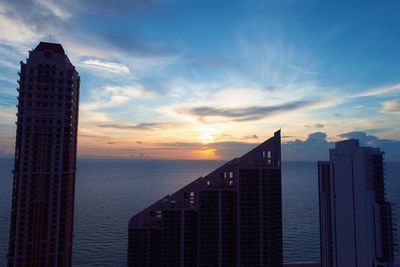 Sea by modern buildings against sky during sunset