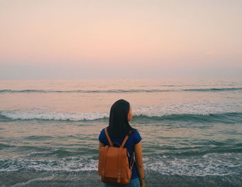 Rear view of woman standing on shore at beach against sky during sunset