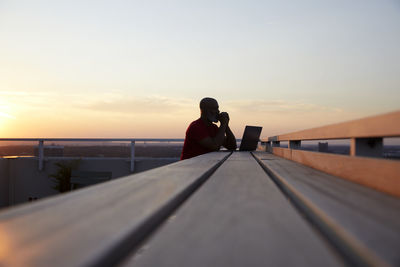 Surface level view of wooden table with man and laptop at rooftop against sky during sunset