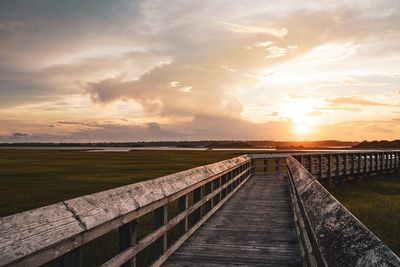 Boardwalk amidst field against sky during sunset