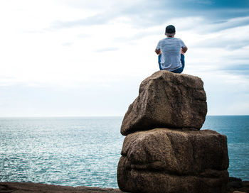 Rear view of man siting on rock by sea against sky