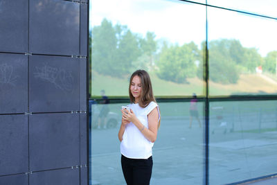 Young businesswoman using phone while standing against wall