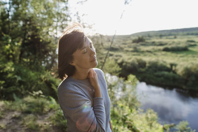 Woman with eyes closed standing at riverbank on sunny day