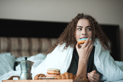 Woman with curly hairstyle eat breakfast in bed with coffee and donuts with white duvet close up.