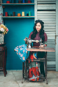 Young woman using sewing machine while sitting against wall