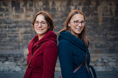 Portrait of smiling sisters standing back to back against stone wall