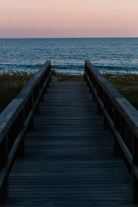 Wooden boardwalk leading to sea against sky during sunset