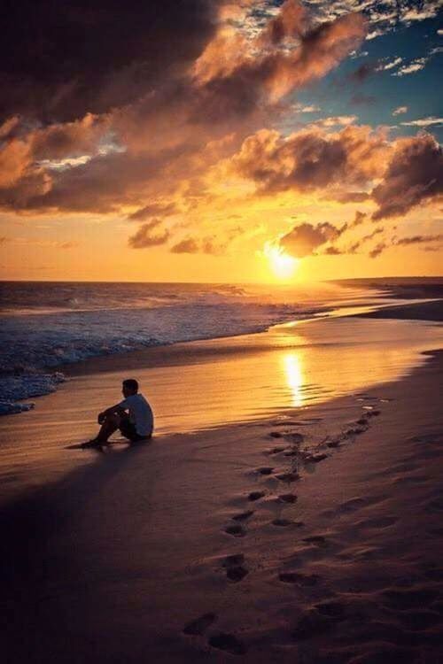 MAN SITTING ON BEACH AGAINST SKY DURING SUNSET