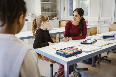 Smiling female teacher and schoolgirl interacting with each other while sitting in classroom