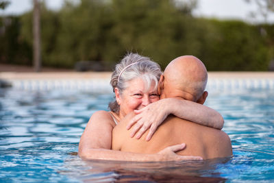 Smiling gray haired woman embracing bald shirtless man while swimming in clean pool water together